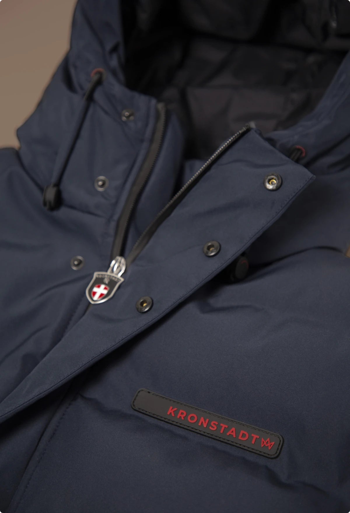 Detailaufnahme einer dunkelblauen Winterjacke von KRONSTADT mit Reißverschluss, Knöpfen und Kapuze mit Kordel, schwarze Innenseite und sichtbares Logo mit rotem Schriftzug