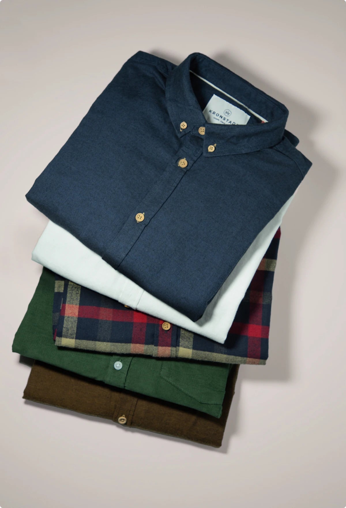 Gestapelte Hemden von KRONSTADT in Braun, Grün, rot-blau-gelb kariert, Weiß und Blau, vor grauem Hintergrund