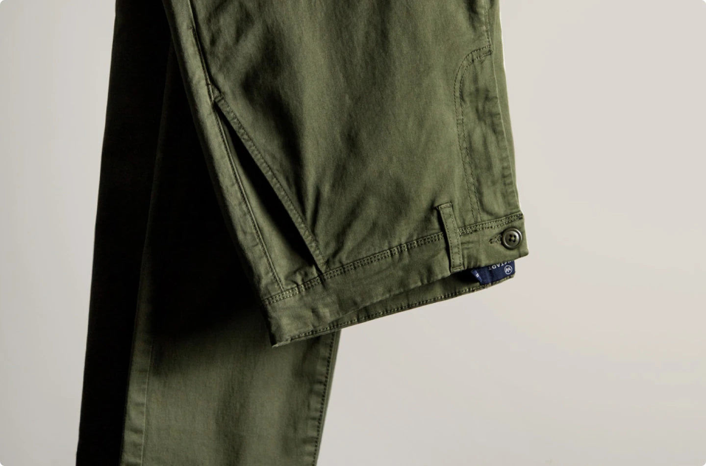 Khakigrüne Hose von KRONSTADT mit Gürtelschlaufen, Knopf und Hosentasche, hängend vor grauem Hintergrund