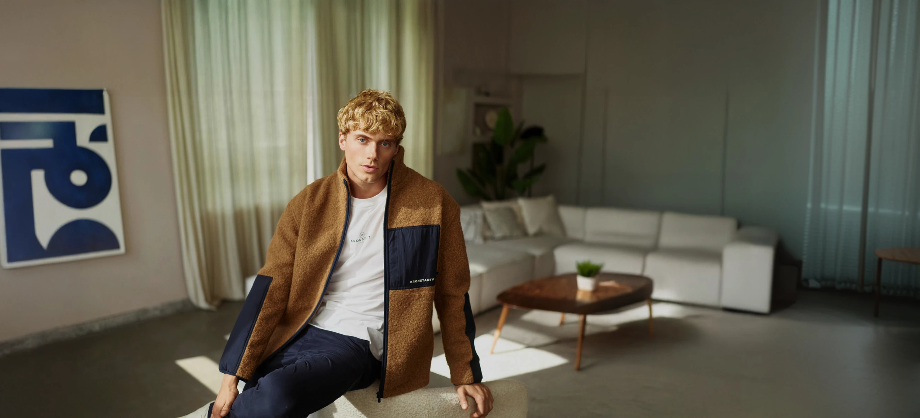 Blondes Model trägt weißes Shirt und Teddyfelljacke mit blauen Akzenten sowie blaue Hose von KRONSTADT, in modernem Wohnzimmer mit Sofa und Holztisch