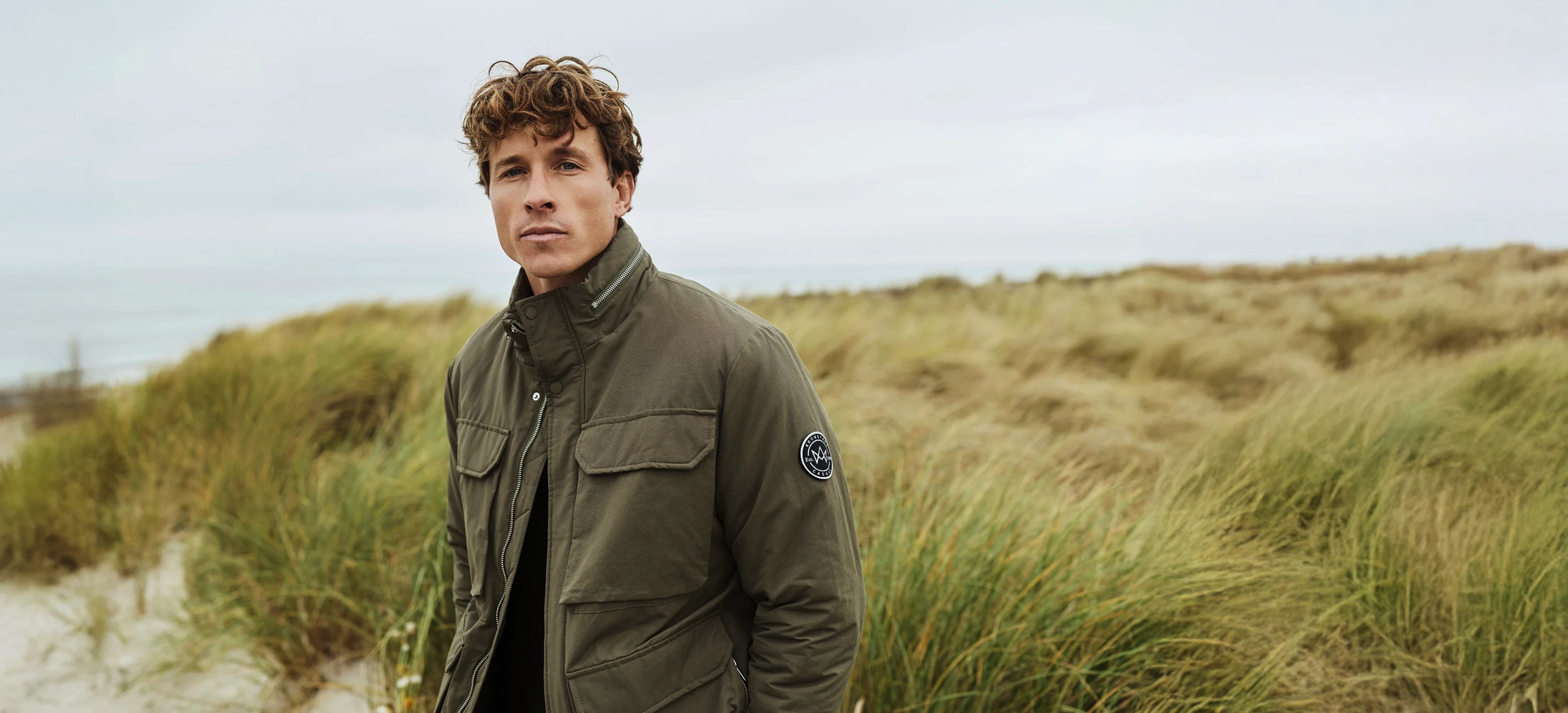 Khakigrüne Übergangsjacke von KRONSTADT mit Brusttaschen und hohem Kragen, getragen von blondem Model in Dünenlandschaft