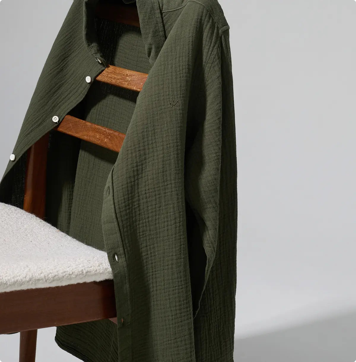 Dunkelgrünes strukturiertes Hemd von KRONSTADT über Holzstuhl mit hellem Sitz gelegt, vor grauem Hintergrund