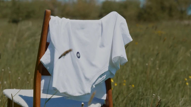 Weißes T-Shirt von KRONSTADT mit rundem schwarzem Logo auf der Brust, über Holzstuhl mit hellem Sitz auf Wiese mit kleinen gelben Blumen
