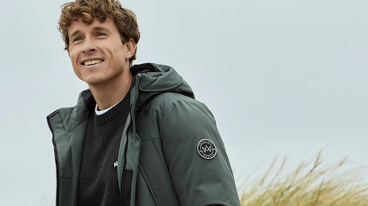 Grüne Kapuzen-Winterjacke von KRONSTADT mit Logo-Patch am Ärmel und dunkelgrauem Pullover mit gesticktem KRONSTADT-Logo, getragen von blondem Model vor Himmel und Dünenlandschaft