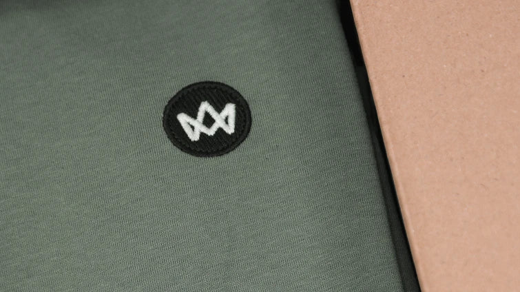 Detailaufnahme eines grünen Kleidungsstücks von KRONSTADT mit gesticktem Logo-Patch in einem Karton