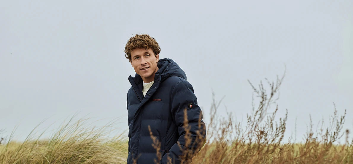 Blaue Winterjacke von KRONSTADT mit rotem Logo, getragen von blondem Model in Graslandschaft unter bewölktem Himmel